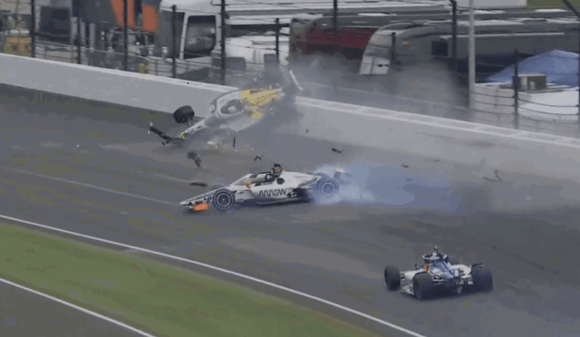 Race car crashes on track, flipping into the air with debris and smoke around the scene at a racing circuit behind a barrier.