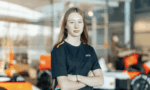 Young woman in a navy uniform with orange accents stands with arms crossed in a bright workshop with orange race cars in the background.