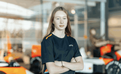 Young woman in a navy uniform with orange accents stands with arms crossed in a bright workshop with orange race cars in the background.