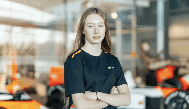 Young woman in a navy uniform with orange accents stands with arms crossed in a bright workshop with orange race cars in the background.