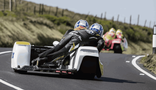 Two riders in matching helmets lean into a left-hand turn on a white sidecar race bike along a rural road.