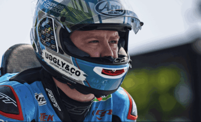 Race car/motorsport rider wearing a blue helmet with sponsor logos (UGLY&CO) and a blue racing suit, looking ahead in focus.