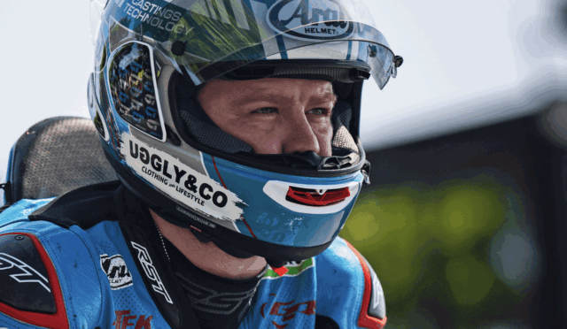 Race car/motorsport rider wearing a blue helmet with sponsor logos (UGLY&CO) and a blue racing suit, looking ahead in focus.