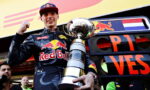 Racing driver in Red Bull suit raising fist with a large trophy in front of a team truck.