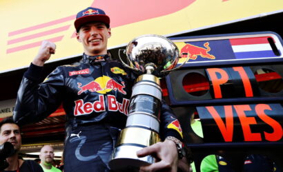 Racing driver in Red Bull suit raising fist with a large trophy in front of a team truck.
