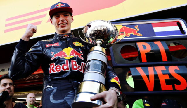 Racing driver in Red Bull suit raising fist with a large trophy in front of a team truck.