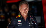 Portrait of a middle-aged man in a navy racing team shirt with sponsor logos (TAG Heuer, Mobil 1, Oracle) in a garage, looking to the side.