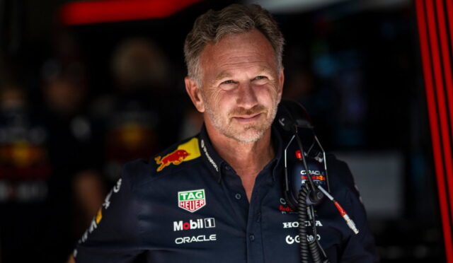 Portrait of a middle-aged man in a navy racing team shirt with sponsor logos (TAG Heuer, Mobil 1, Oracle) in a garage, looking to the side.