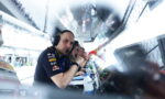 Pit crew member with headset looking at the camera in a race garage, with monitors and teammates in the background.