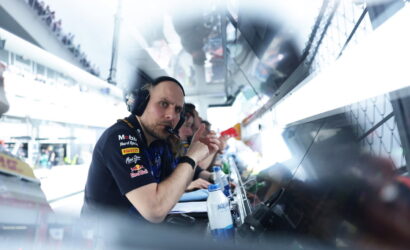 Pit crew member with headset looking at the camera in a race garage, with monitors and teammates in the background.