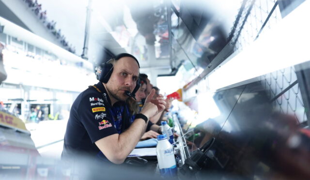 Pit crew member with headset looking at the camera in a race garage, with monitors and teammates in the background.