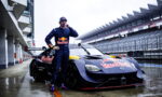 Race car driver in a Red Bull suit stands beside a wet purple Nissan race car in the pit lane, drinking from a bottle.