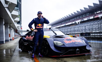 Race car driver in a Red Bull suit stands beside a wet purple Nissan race car in the pit lane, drinking from a bottle.