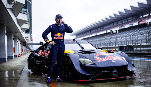 Race car driver in a Red Bull suit stands beside a wet purple Nissan race car in the pit lane, drinking from a bottle.