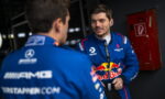 Two Formula 1 drivers in blue Red Bull racing suits chat in a dim pit area, one wearing earphones and smiling at the other.