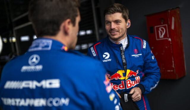 Two Formula 1 drivers in blue Red Bull racing suits chat in a dim pit area, one wearing earphones and smiling at the other.