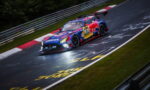Blue and red racing car with sponsor logos speeding through a corner on a wet race track, guard rails in the background and motion blur indicating high speed.