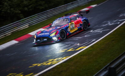 Blue and red racing car with sponsor logos speeding through a corner on a wet race track, guard rails in the background and motion blur indicating high speed.