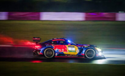 Blue race car with red and yellow sponsor graphics, number 3, speeding at night on a track with bright headlights and motion blur behind it.