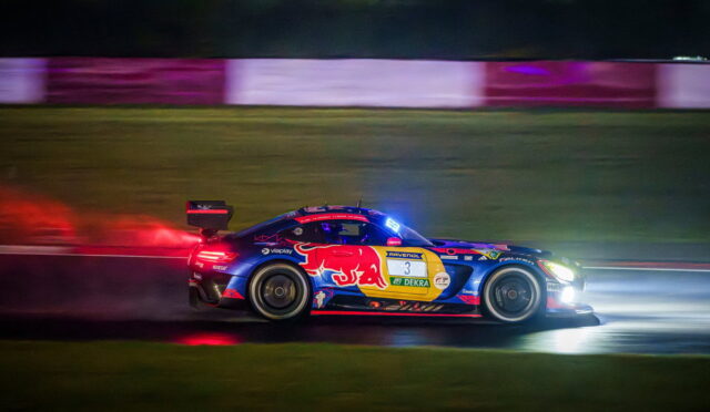 Blue race car with red and yellow sponsor graphics, number 3, speeding at night on a track with bright headlights and motion blur behind it.