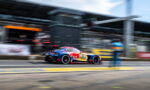 Blue-and-red race car with sponsor decals speeds along the pit lane, background blurred by motion.