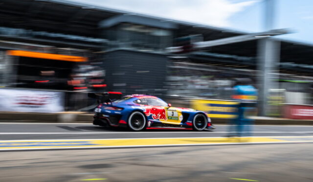 Blue-and-red race car with sponsor decals speeds along the pit lane, background blurred by motion.
