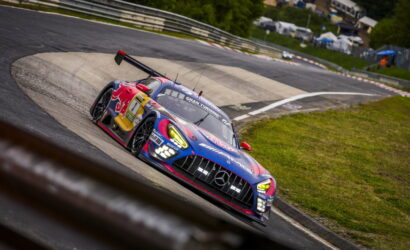 Blue and red race car (Mercedes) leans into a sharp turn on a race track with tire marks and grassy infield in the background.