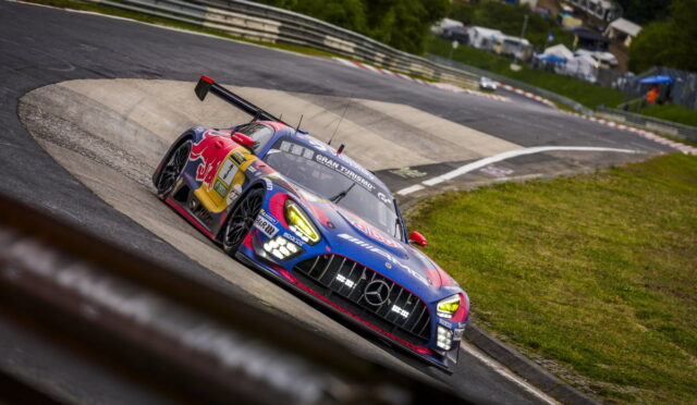 Blue and red race car (Mercedes) leans into a sharp turn on a race track with tire marks and grassy infield in the background.