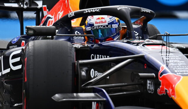 Formula 1 race car close-up: cockpit with Red Bull branding, helmeted driver visible behind the steering wheel, sponsor logos around the fuselage.