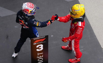 Two Formula 1 drivers in racing suits shake hands on a podium next to a Moët & Chandon sign with a '3' marker, viewed from above on a dark-gray podium surface.