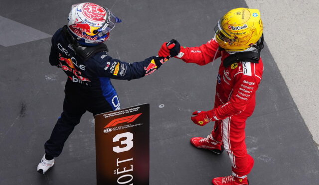 Two Formula 1 drivers in racing suits shake hands on a podium next to a Moët & Chandon sign with a '3' marker, viewed from above on a dark-gray podium surface.