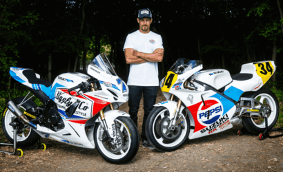 Man in a white shirt stands with two white Suzuki racing motorcycles on a dirt path in a forest clearing.