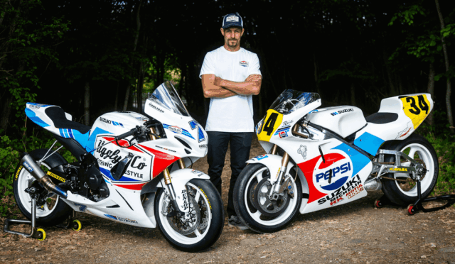 Man in a white shirt stands with two white Suzuki racing motorcycles on a dirt path in a forest clearing.