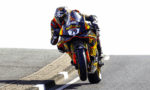 Sport motorcycle racer in full leathers leans into a left-hand bend, front wheel lifted off the road during a high-speed turn on a track-like roadway.