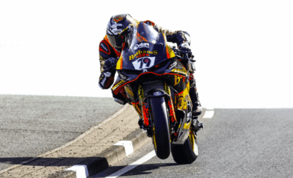 Sport motorcycle racer in full leathers leans into a left-hand bend, front wheel lifted off the road during a high-speed turn on a track-like roadway.
