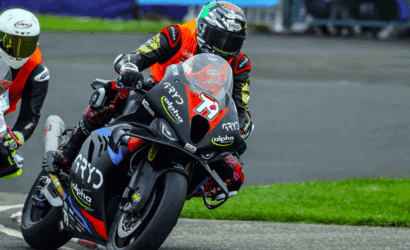 Two motorcycle racers lean into a turn on a race track, foreground rider on a black and red bike (#15) wearing orange and black gear with sponsor logos: Alpha Racing, GPY, and SHOE. Behind them, another rider in a white helmet follows on a white bike.