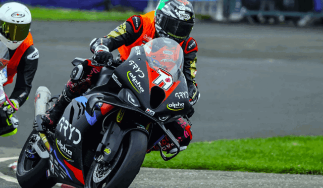 Two motorcycle racers lean into a turn on a race track, foreground rider on a black and red bike (#15) wearing orange and black gear with sponsor logos: Alpha Racing, GPY, and SHOE. Behind them, another rider in a white helmet follows on a white bike.