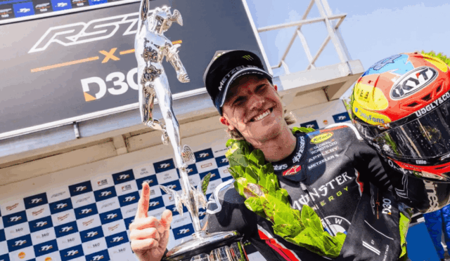 Race winner on podium holding a tall silver trophy, wearing a racing suit and helmet, with a laurel wreath draped over the shoulder.