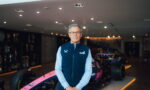 A man in a blue quilted vest and glasses stands beside a pink race car in a showroom.