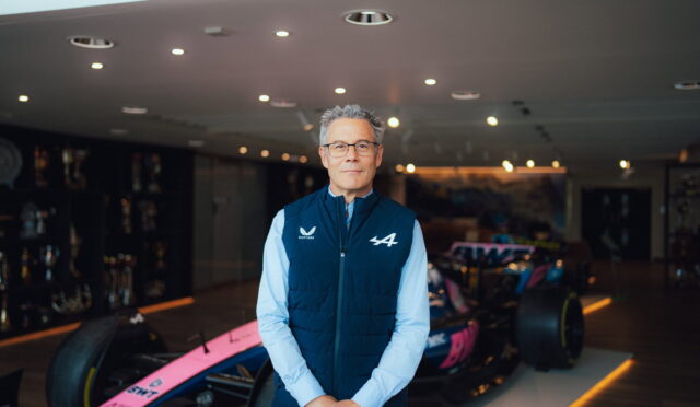 A man in a blue quilted vest and glasses stands beside a pink race car in a showroom.