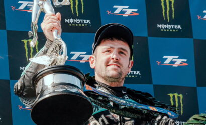 Man in racing gear on a winner's podium lifts a silver trophy against a Monster Energy backdrop at a blue checkered stagework backdrop.