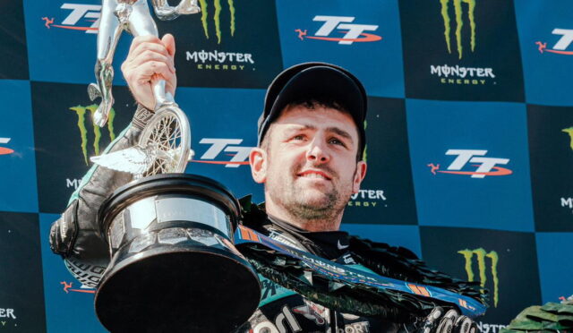 Man in racing gear on a winner's podium lifts a silver trophy against a Monster Energy backdrop at a blue checkered stagework backdrop.