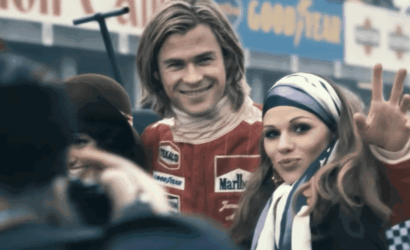 Smiling race car driver in a red suit poses with a fan wearing a striped headscarf at a race event, giving a peace sign to the camera.