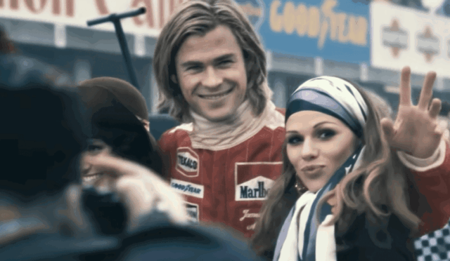 Smiling race car driver in a red suit poses with a fan wearing a striped headscarf at a race event, giving a peace sign to the camera.