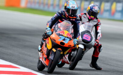 Two MotoGP riders lean into a turn on a race track; leading rider wears orange KTM with #73 and Red Bull branding, followed by a white/grey bike with a bold purple logo guide bike