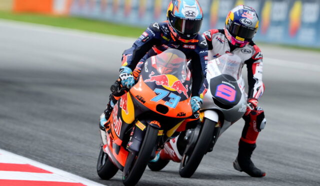 Two MotoGP riders lean into a turn on a race track; leading rider wears orange KTM with #73 and Red Bull branding, followed by a white/grey bike with a bold purple logo guide bike