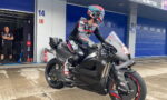 Race rider in full leathers and helmet sits astride a matte black sport motorcycle in a pit area with blue garage doors in the background, ready for practice.