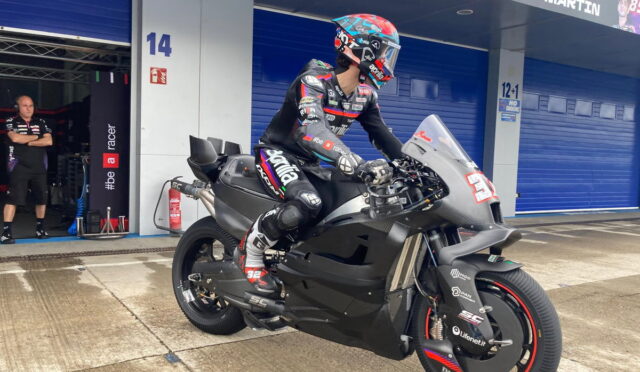 Race rider in full leathers and helmet sits astride a matte black sport motorcycle in a pit area with blue garage doors in the background, ready for practice.