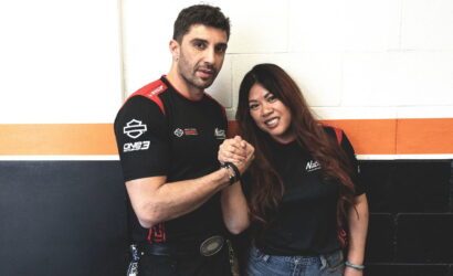 Two teammates in black and red shirts shake hands and smile against a white wall with orange and black stripes behind them.