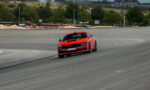 Red sports car drifting around a race track, tire marks visible on the asphalt with green trees in the background.